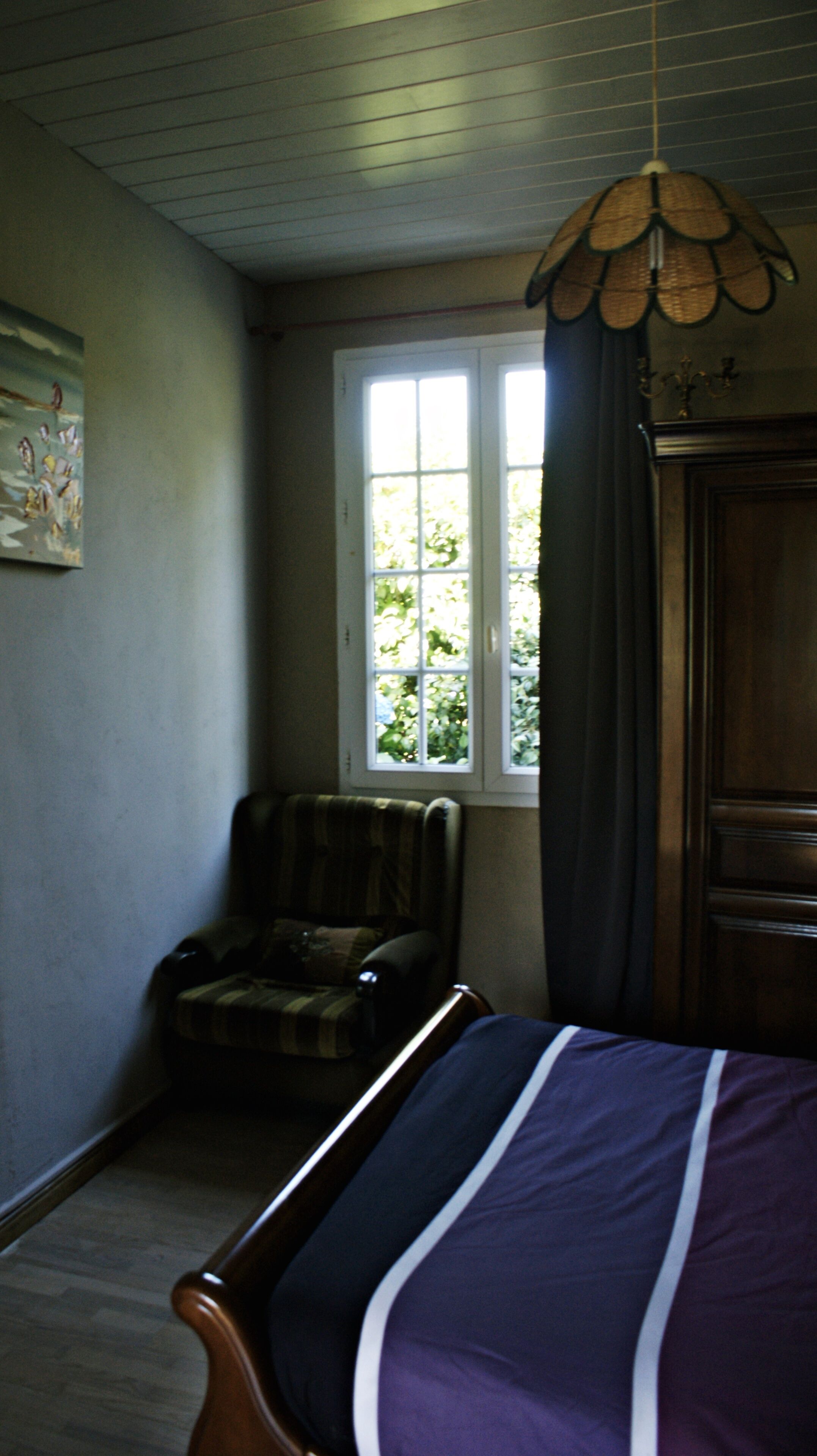 A bedroom with a bed, a chair, a window with curtains, and a hanging light fixture.