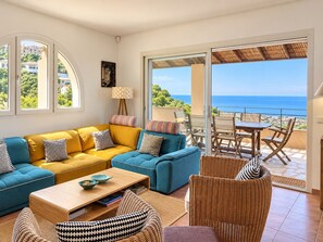 A living room with a sofa, coffee table, and wicker chairs, a balcony with a table and chairs, and a view of the sea.