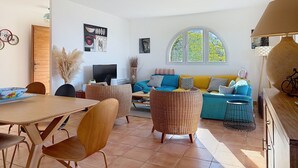 Amplia sala de estar con un sofá grande, una mesa de comedor con sillas, un televisor y una ventana con vista a la vegetación.