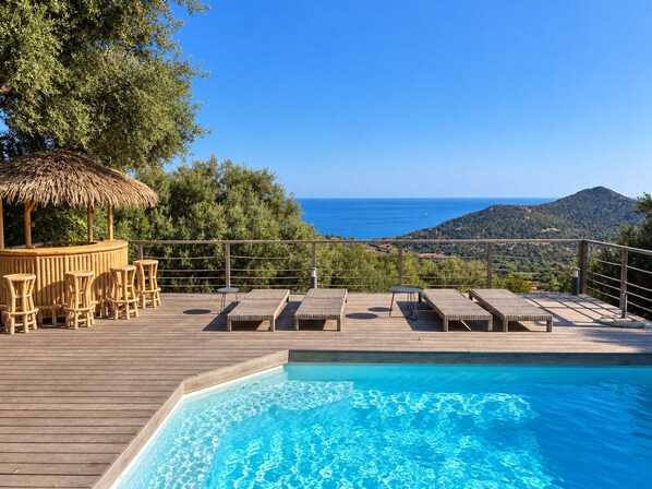 A wooden deck with a pool, lounge chairs, and a bar area overlooking a mountainous landscape and the sea.