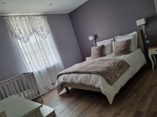 A bedroom with a bed, a bedside table, a lamp, a crib, and a window with sheer curtains.