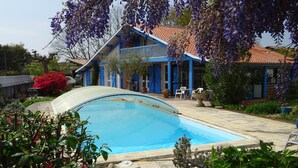 Une piscine entourée d’une végétation luxuriante et une maison à l’extérieur bleue et au toit recouvert de tuiles rouges.
