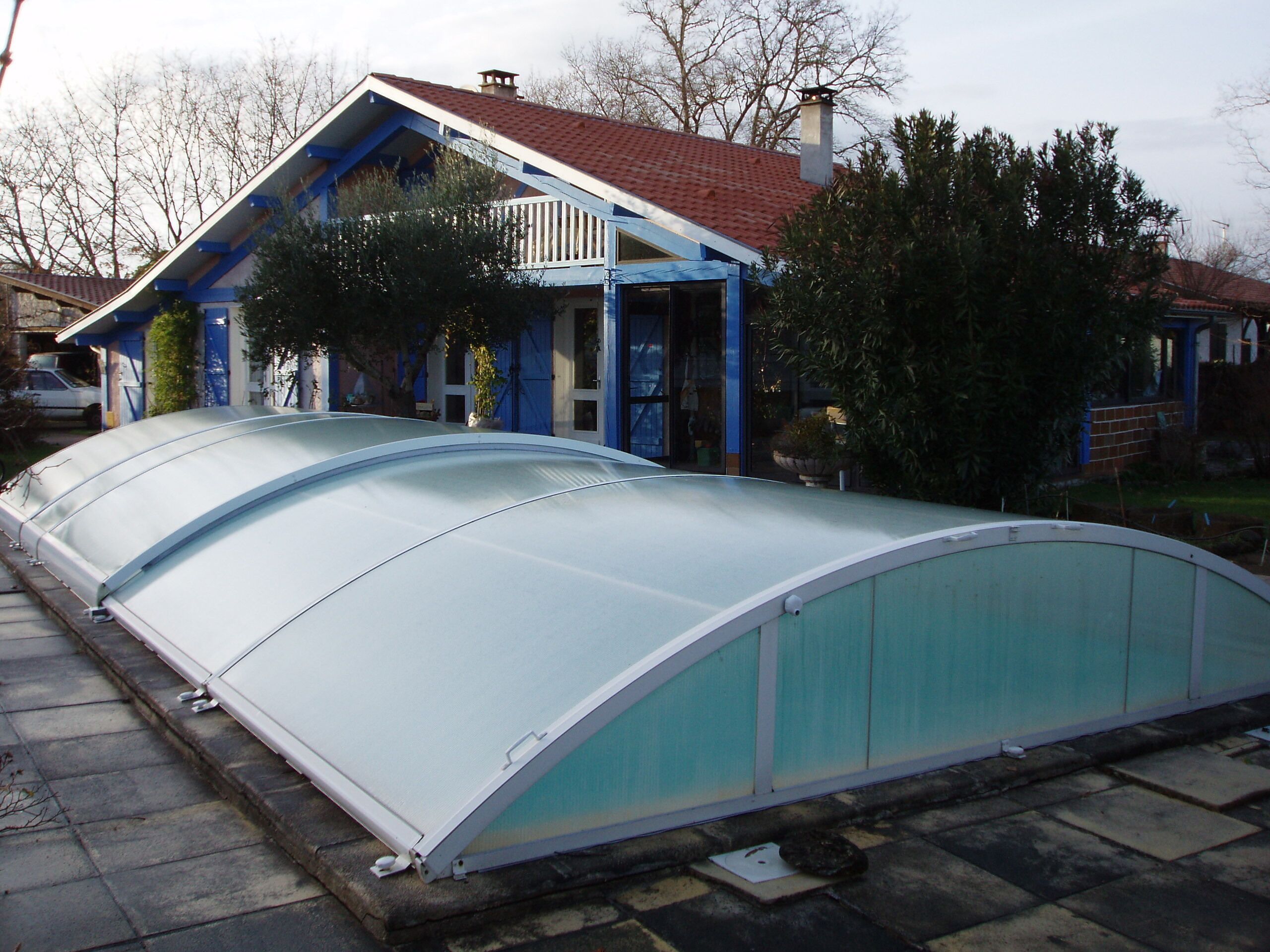 En växthuskonstruktion med ett böjt tak, placerad intill ett hus med rött tak och blåa detaljer.