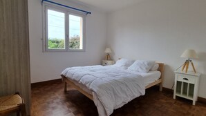 Un dormitorio con una cama de madera, una mesita de noche pequeña con una lámpara, una ventana con persiana azul y una silla.