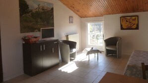 A room with a wooden ceiling, a painting of a landscape, a television on a cabinet, a chair, a small table with a cup, and a window with a view of trees. - furnished studio overlooking the luberon (Lagnes)