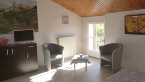 A room with a wooden ceiling, a painting of a village, a TV on a cabinet, two chairs, a small table with a bowl, and a window with a view of greenery. - furnished studio overlooking the luberon (Lagnes)