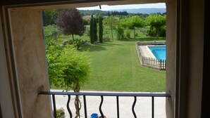 A balcony with a view of a backyard featuring a swimming pool, green lawn, and trees. - furnished studio overlooking the luberon (Lagnes)