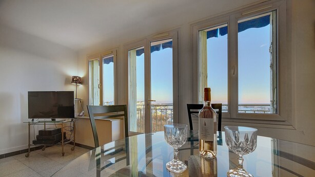 A dining area with a glass table, chairs, a bottle of wine, and a TV on a stand.
