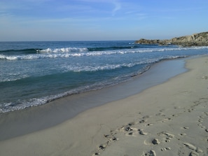 A sandy beach with footprints leading to the water, a rocky outcrop in the distance, and waves gently crashing onto the shore. - 2-room apartment, new with terrace and sea view sleeping 2 to 4 (L'île-Rousse)