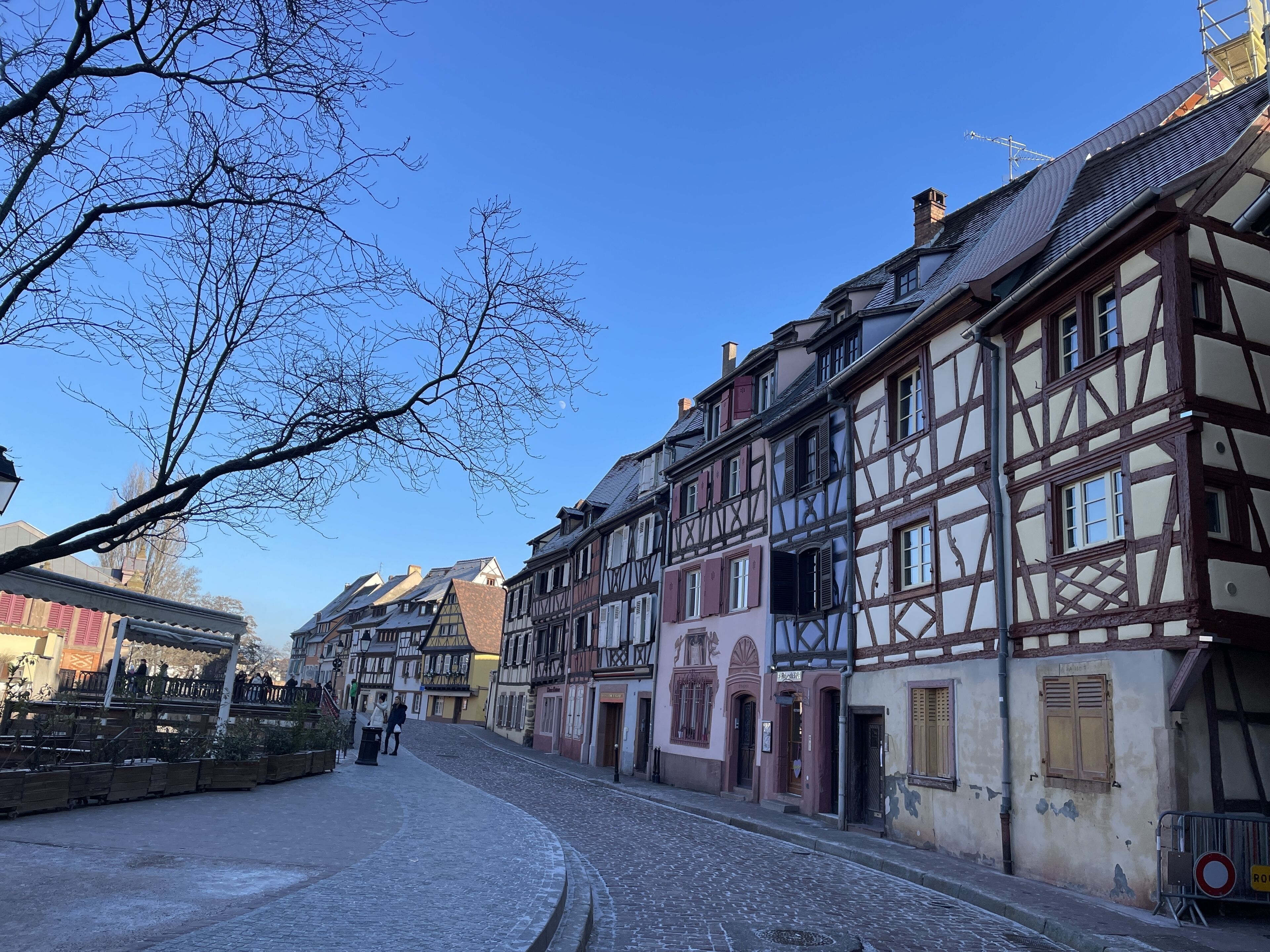 Une rue pavée bordée de bâtiments traditionnels à colombages, un ciel d’un bleu limpide et quelques piétons.