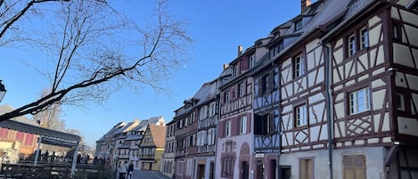Une rue pavée bordée de bâtiments traditionnels à colombages, un ciel d’un bleu limpide et quelques piétons.