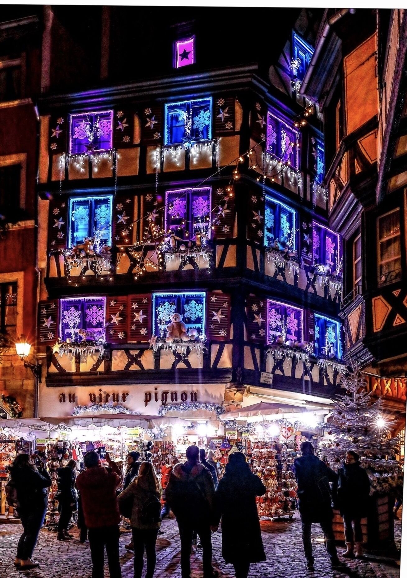 A traditional European building with multiple illuminated windows and balconies, surrounded by a bustling market area.