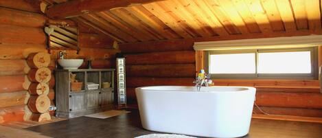 A log cabin bathroom with a large bathtub, wooden walls, and a window providing natural light.