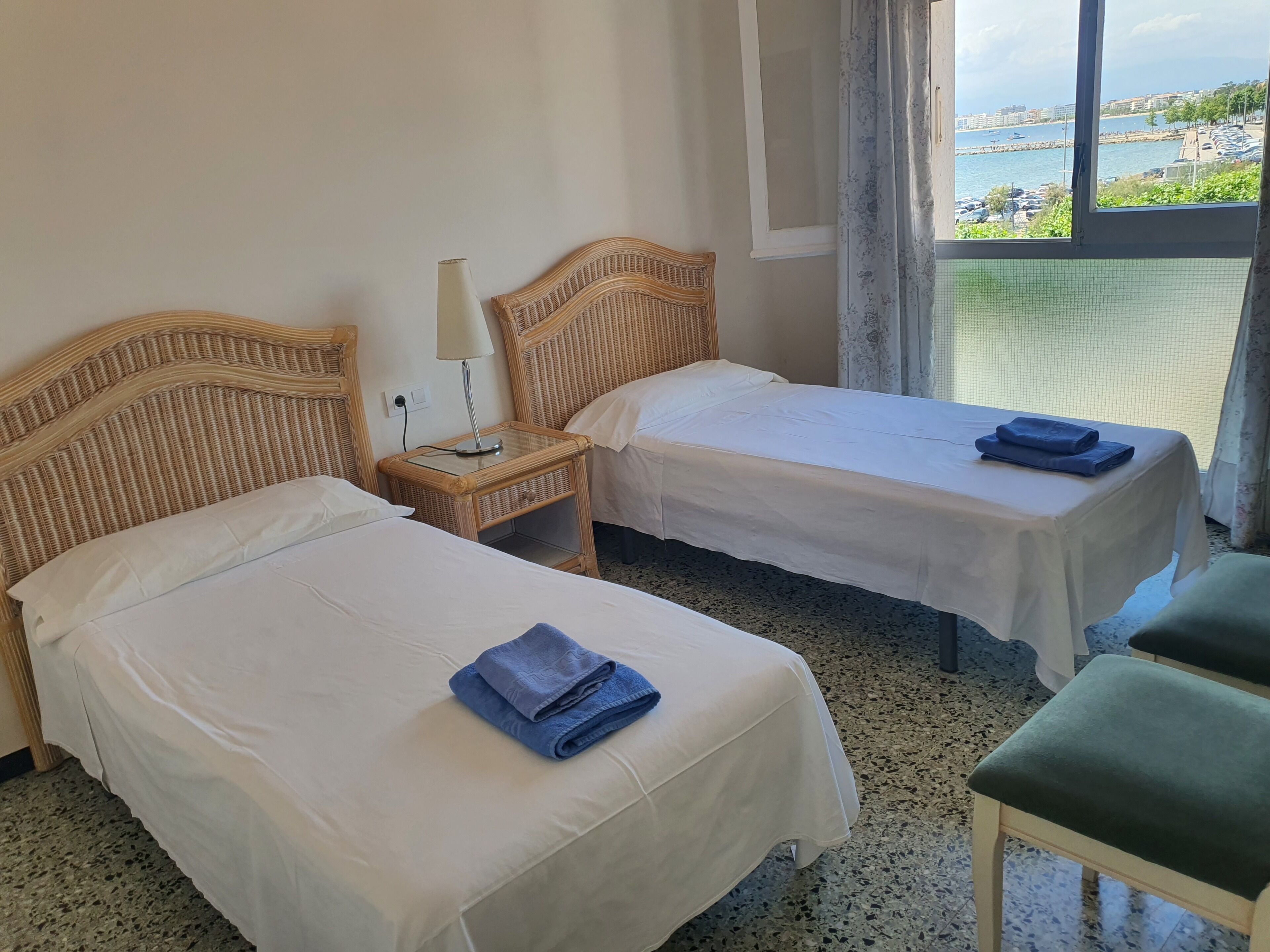 Habitación de hotel con dos camas individuales, una mesita con lámpara y vista al mar a través de una ventana.