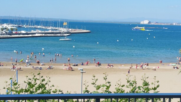 Una playa con gente tomando el sol y nadando, una marina con barcos, y un paisaje urbano al fondo.
