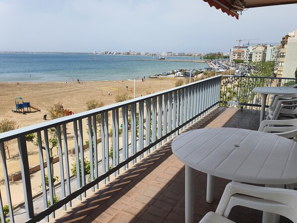 A balcony with a view of a beach, a parking area, and a row of white plastic chairs. - NICE APARTMENT IN FRONT OF THE BEACH FOR 8 PEOPLE IN THE CENTER OF ROSES (Roses)