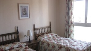 Un dormitorio con dos camas, una mesita de noche con lámpara, un cuadro floral en la pared y una ventana con cortinas de motivos florales.