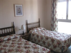 A bedroom with two beds, a nightstand with a lamp, a floral painting on the wall, and a window with floral curtains.