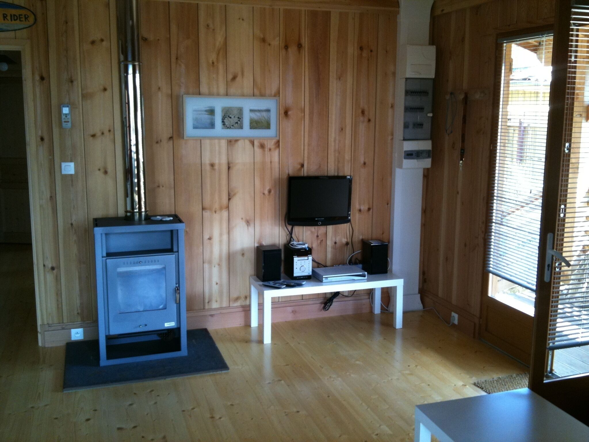 Quarto com paredes de madeira, uma televisão em um suporte, uma pequena mesa com dispositivos eletrônicos e uma porta com janela.
