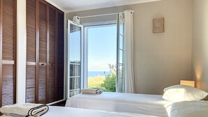 Quarto com duas camas, uma janela grande com vista para o verde e o mar, e um guarda-roupa de madeira.