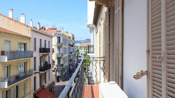 Una calle estrecha flanqueada por edificios, un balcón con pared blanca y contraventanas, y un toldo rojo en una de las construcciones.