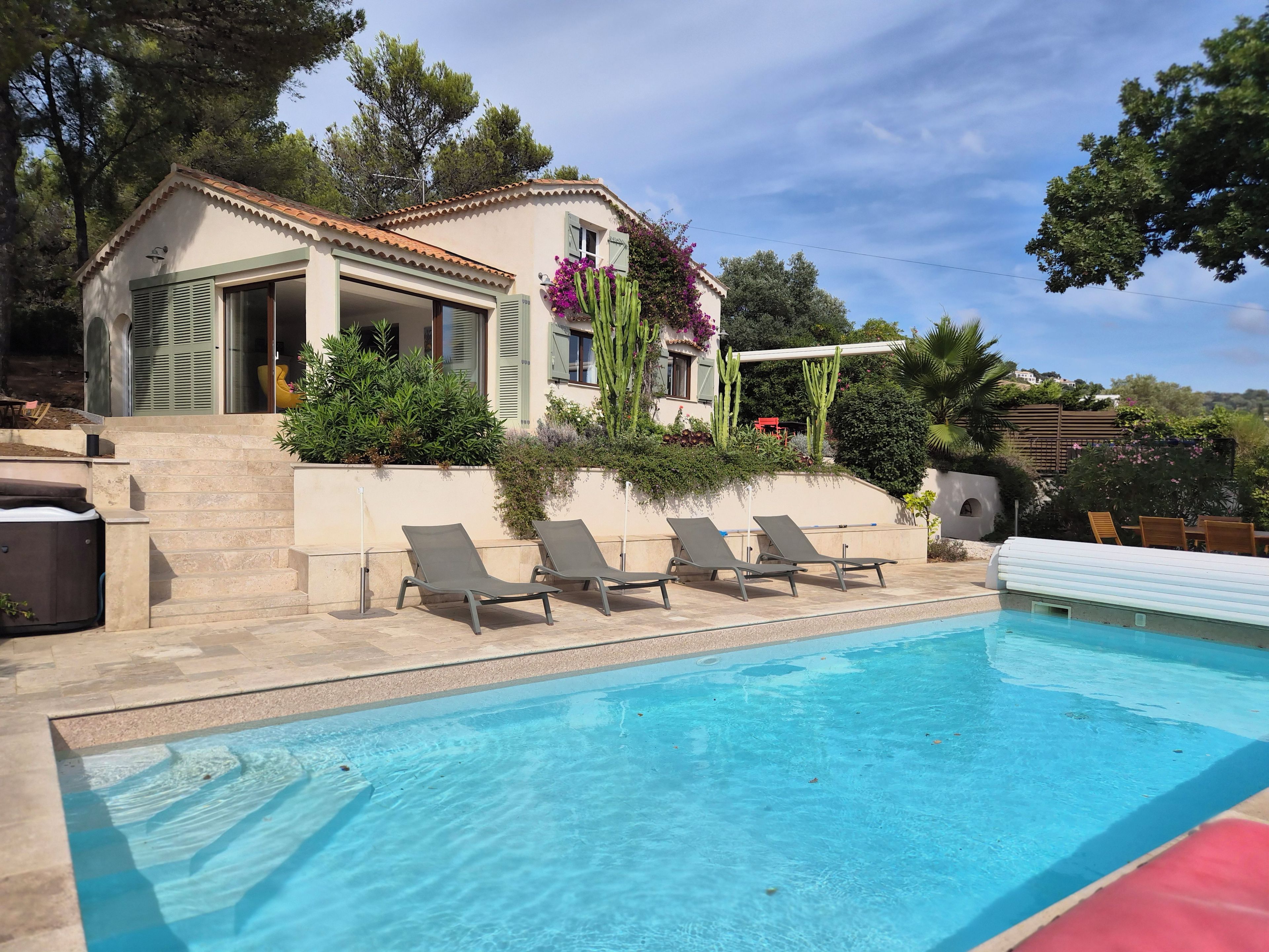 A pool with lounge chairs, a house with a tiled roof, and surrounding greenery.
