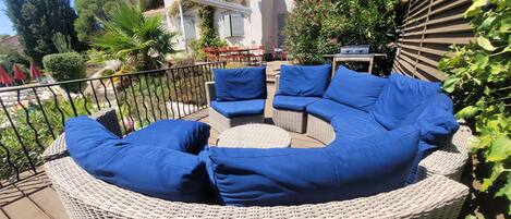 A wicker outdoor seating area with blue cushions, a wooden table, and a house with a balcony in the background.