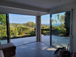 Ein geräumiger Raum mit großen Schiebeglastüren, die auf eine Terrasse mit Blick auf eine üppige Landschaft und das Meer führen.