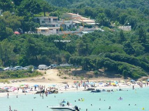 La villa Angelique est entourée d’une végétation luxuriante, et on peut y admirer une plage animée, avec des gens et des bateaux, sous un ciel dégagé.