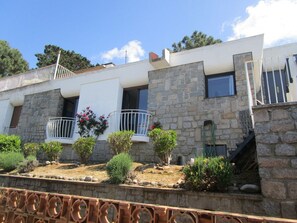 Une maison à deux étages avec une façade en pierre, un balcon doté d’une balustrade blanche et un petit jardin agrémenté de diverses plantes.