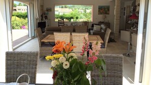 A spacious living area with wicker furniture, a large window offering a view of greenery, and a dining table with a vase of flowers. - Provencal bastide with heated pool and air conditioning in Plascassier (Mouans-Sartoux)