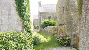 Un étroit sentier de pierre, bordé de murs de pierre et d’une végétation luxuriante, mène à un bâtiment rustique en pierre.