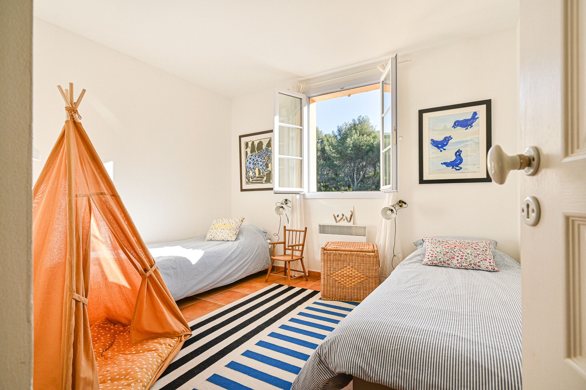 A bedroom with two beds, a window, a chair, a lamp, and a woven basket.