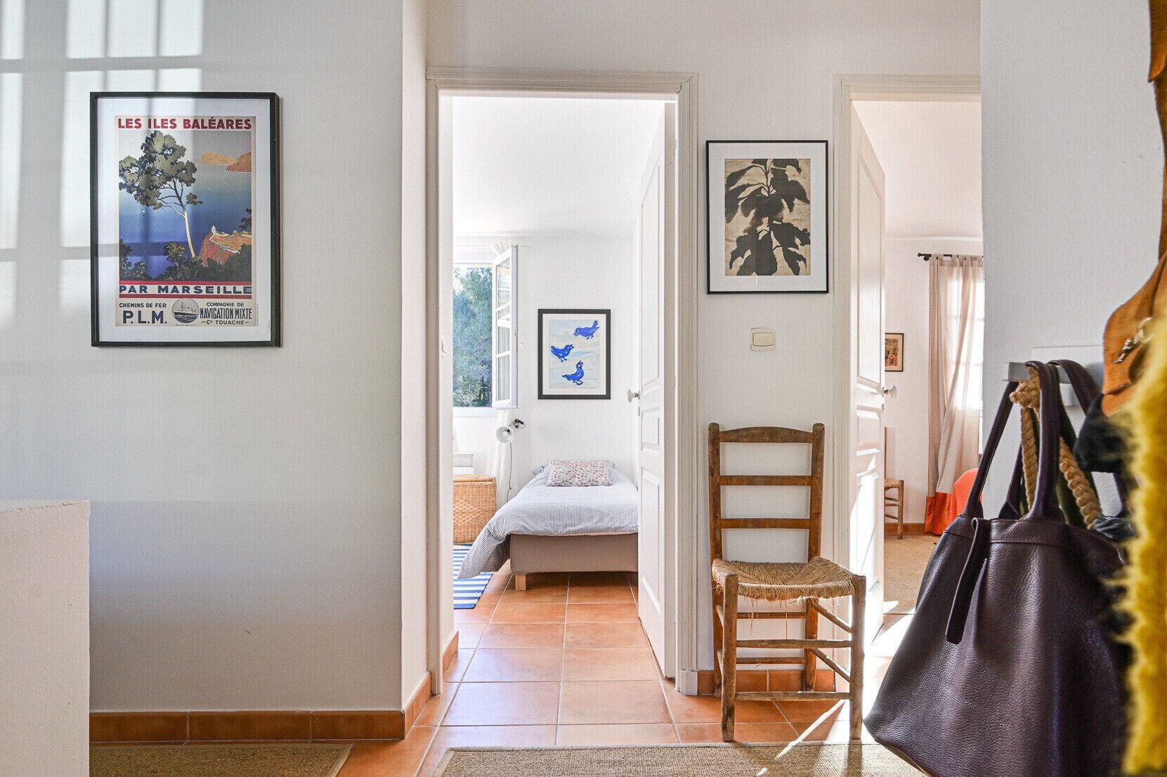 Un pasillo que conduce a un dormitorio con una cama, una silla y una puerta. Hay cuadros enmarcados en las paredes.