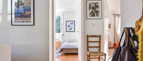 Un pasillo que conduce a un dormitorio con una cama, una silla y una puerta. Hay cuadros enmarcados en las paredes.