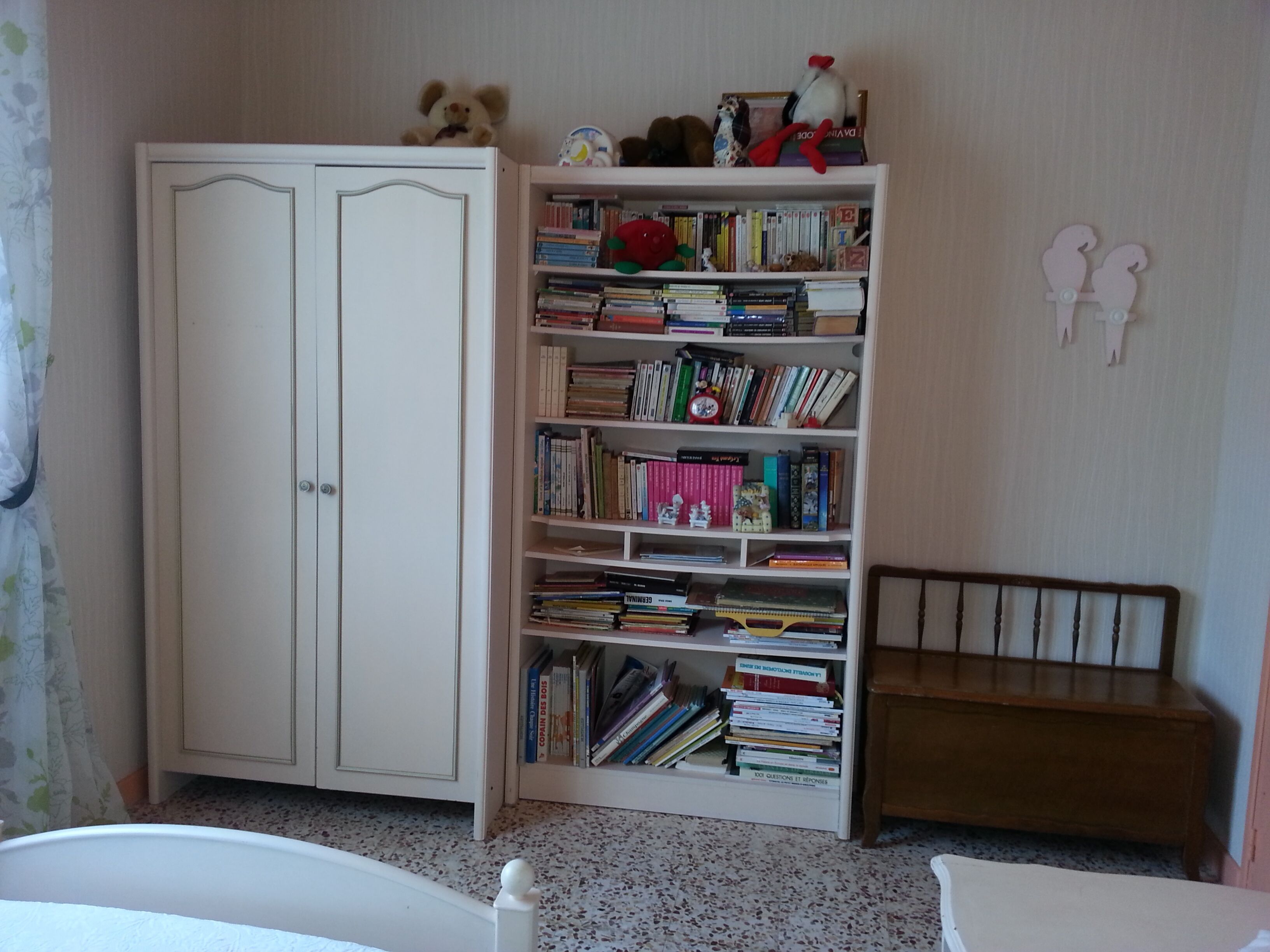 Una habitación con un armario blanco, una estantería llena de libros y juguetes, y un banco de madera.