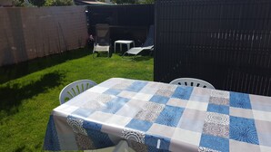 Une table recouverte d’une nappe à carreaux, entourée de chaises blanches, dans un jardin avec une clôture et une petite table ornée de fleurs.