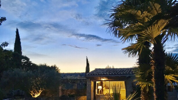 Vue d’une maison avec une façade en pierre, une porte bien visible et un petit patio, le tout vu depuis la piscine.