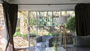 Un salon moderne avec un canapé gris, une table basse blanche et un lampadaire. De grandes fenêtres offrent une vue sur un jardin et un mur en pierre.