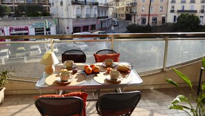 Un balcon avec une table dressée pour le petit-déjeuner, donnant sur une rue bordée d’immeubles et de voitures garées.