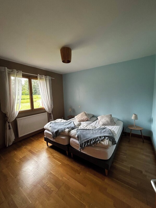 Un dormitorio con cama, ventana con cortinas, radiador y lámpara sobre una mesita de noche.