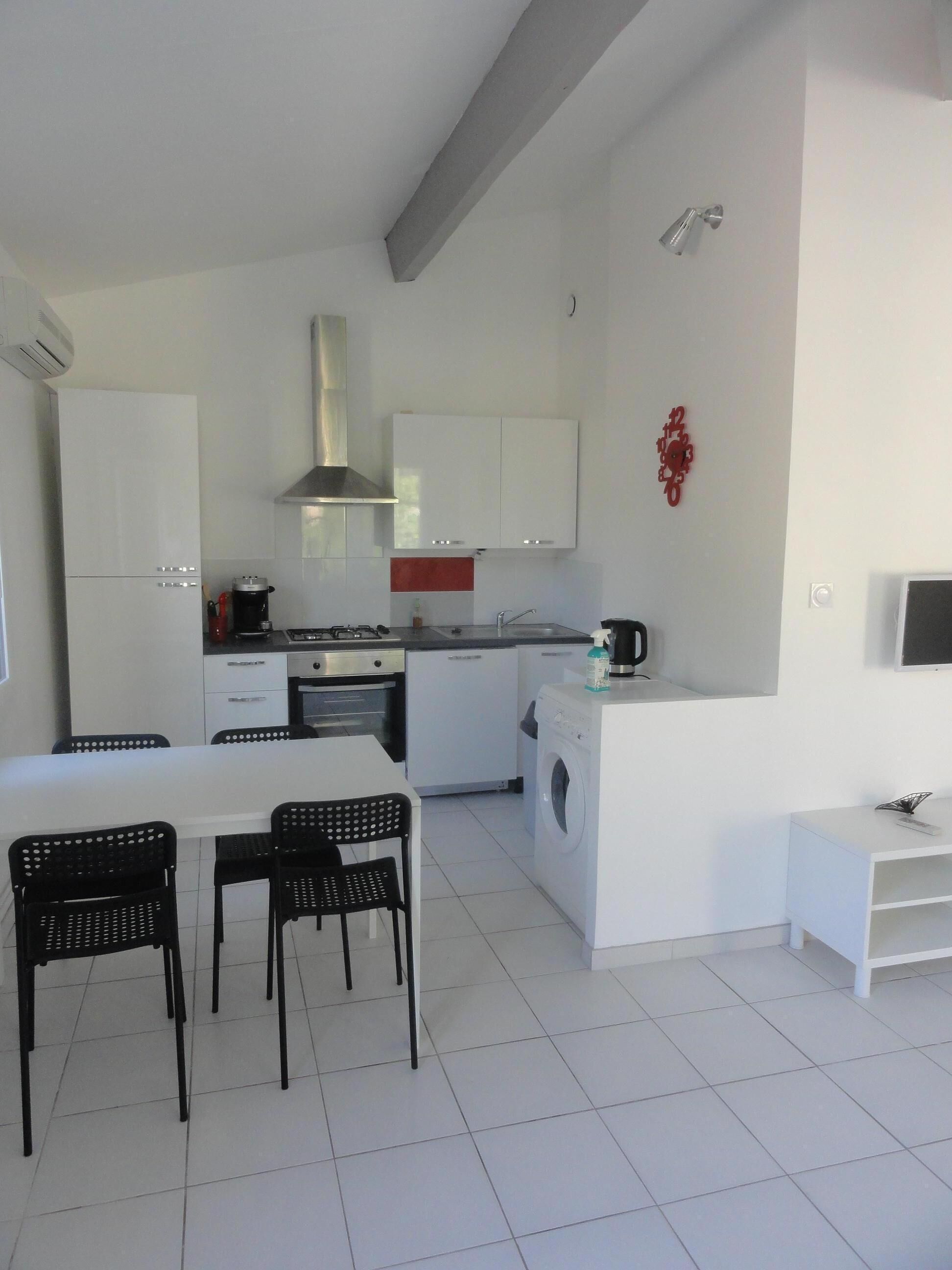 A modern kitchen with white cabinets, a stainless steel range hood, a built-in washing machine, and a dining area with a table and chairs.