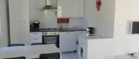 A modern kitchen with white cabinets, a stainless steel range hood, a built-in washing machine, and a dining area with a table and chairs.