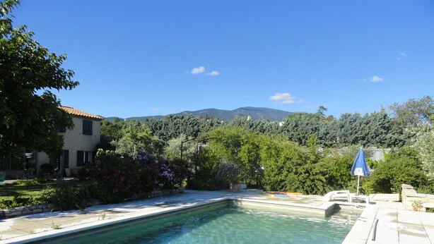 Una piscina cristalina con un borde empinado y azulejos, rodeada de abundante vegetación y una casa con tejado de cerámica.
