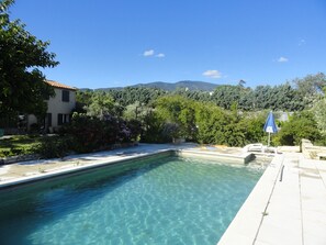Une piscine aux eaux cristallines, bordée d’une terrasse carrelée, entourée d’une végétation luxuriante et d’une maison au toit recouvert de tuiles.
