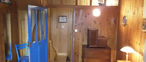 A wooden room with a blue door, a small table with a lamp, a chair, and a cabinet.