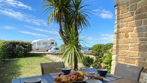 An outdoor patio with a table set for breakfast, surrounded by chairs, a palm tree, and a stone wall. - Loriot1 apartment sea view on the fort blocked, ploemeur  (Ploemeur)