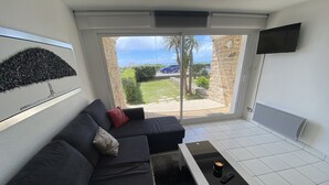 A modern living room with a dark grey sofa, a small black coffee table, and a sliding glass door leading to a balcony with a view of palm trees and a blue car. - Loriot1 apartment sea view on the fort blocked, ploemeur  (Ploemeur)