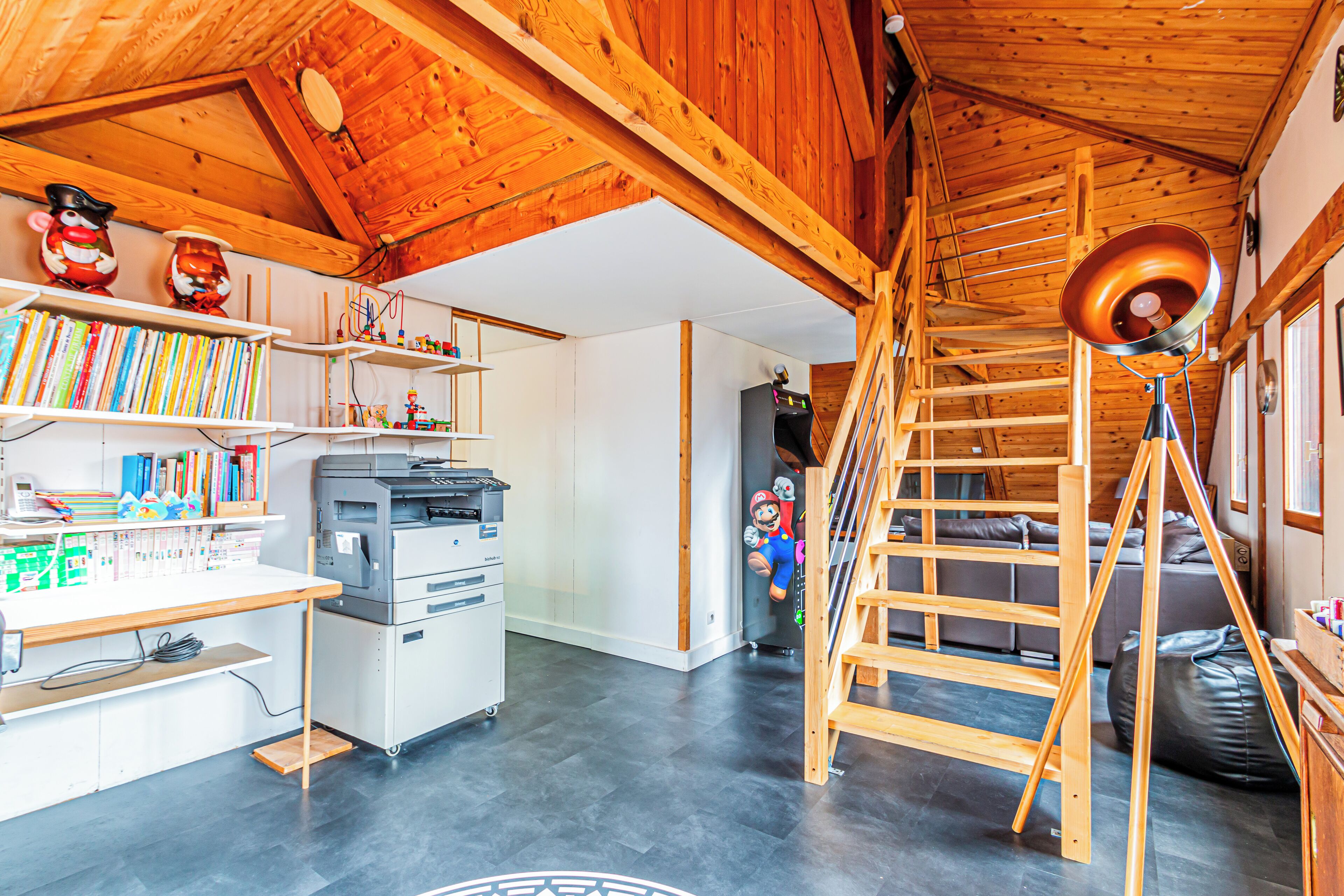 Une pièce avec un plafond en bois, un bureau avec une imprimante, des étagères remplies de livres, une statue de Mario et un fauteuil en forme de poire.