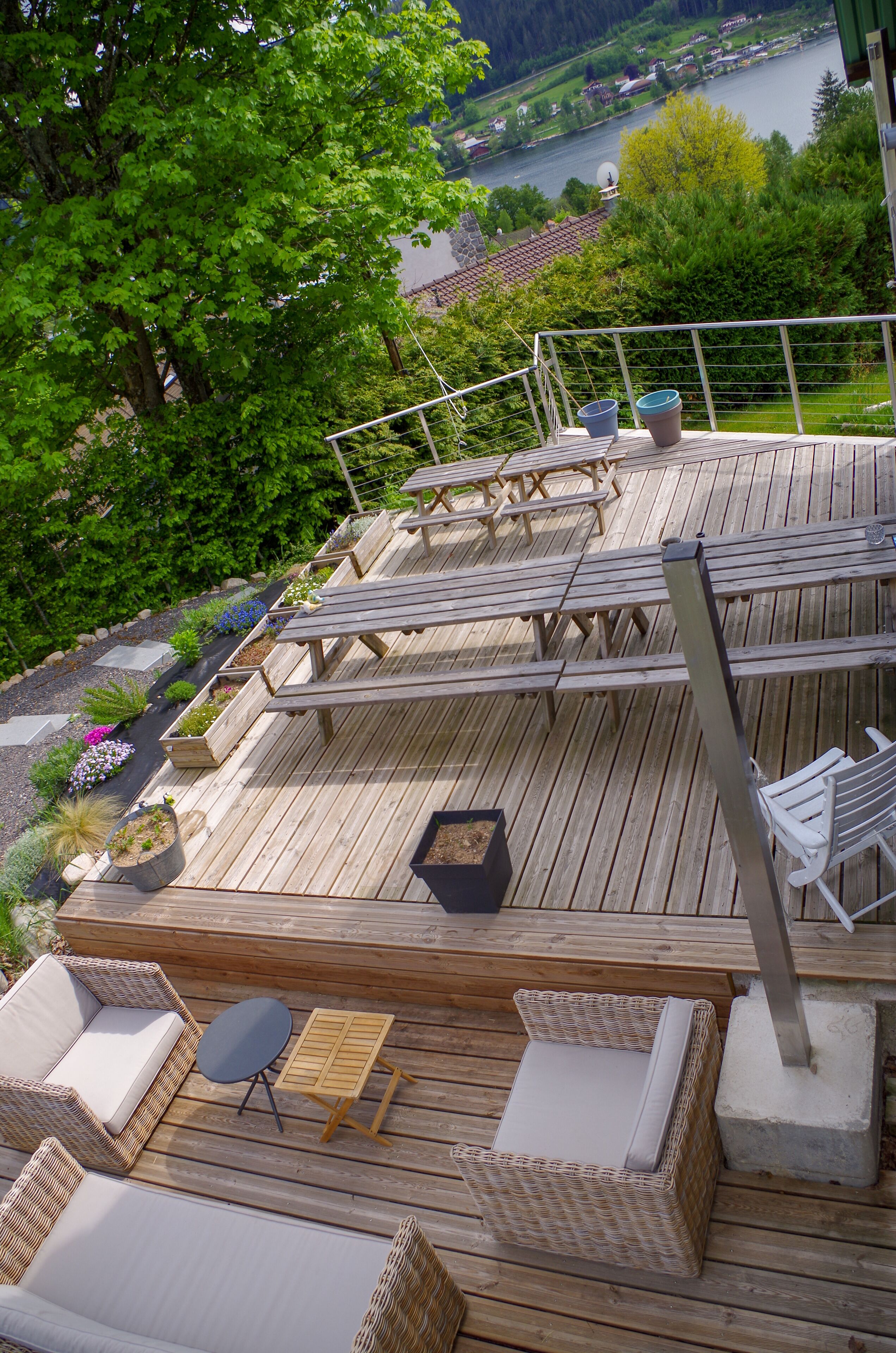 Una terraza de madera con mobiliario exterior, incluyendo sofás de mimbre y una mesa, con vistas a un lago y casas al fondo.